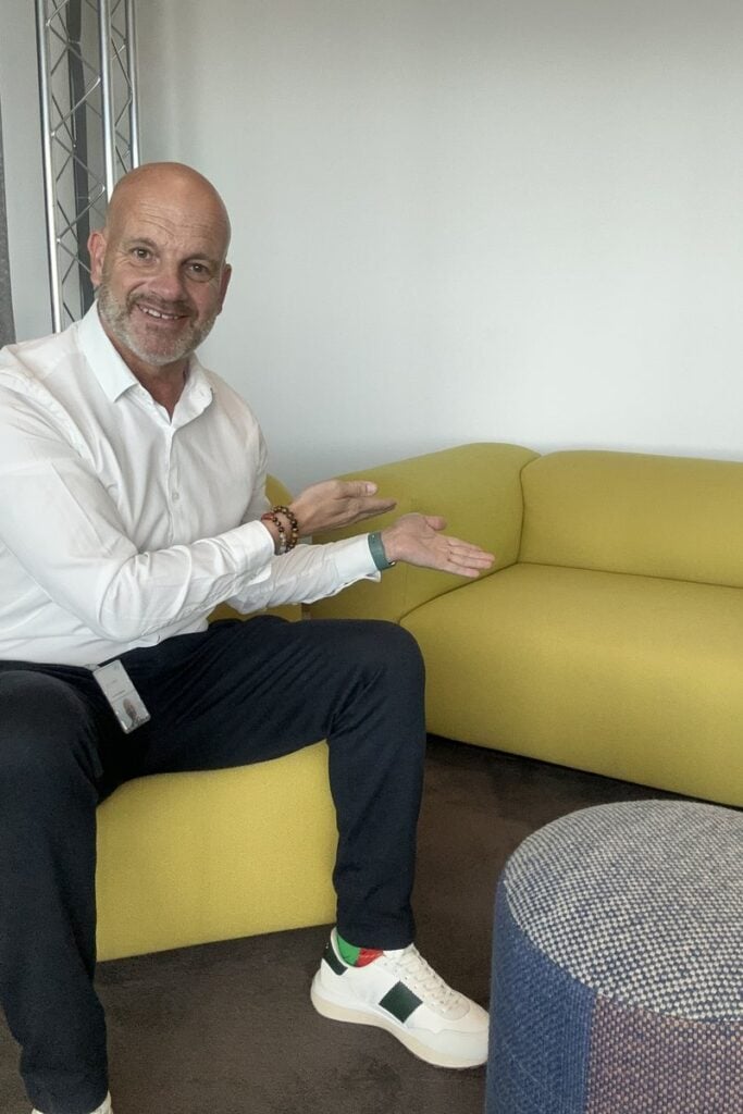 Thorsten Enderlin sitzt auf einem gelben Sofa und lädt mit offener Hand zum Platznehmen ein